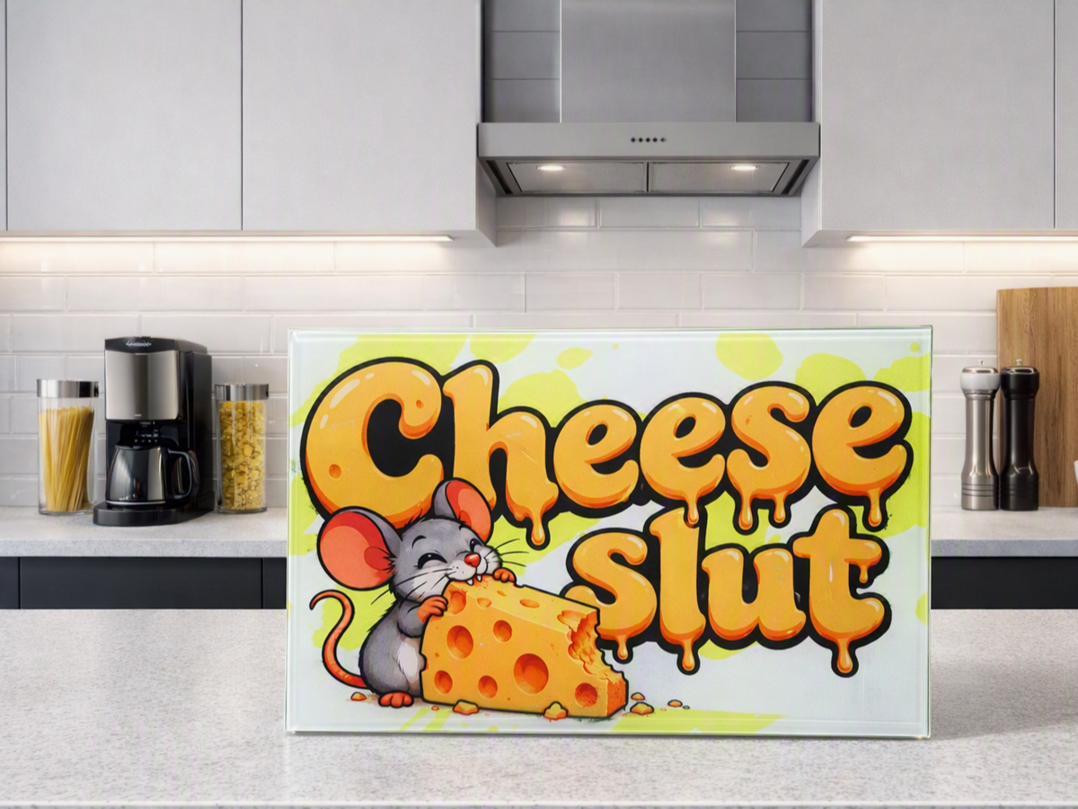 A rectangular kitchen chopping board with a humorous graphic reading 'Cheese slut' in dripping orange-yellow bubble letters, featuring a cartoon grey mouse eating cheese. The chopping board is displayed on a light grey kitchen counter with cabinets, a coffee maker, and other kitchen items in the background.