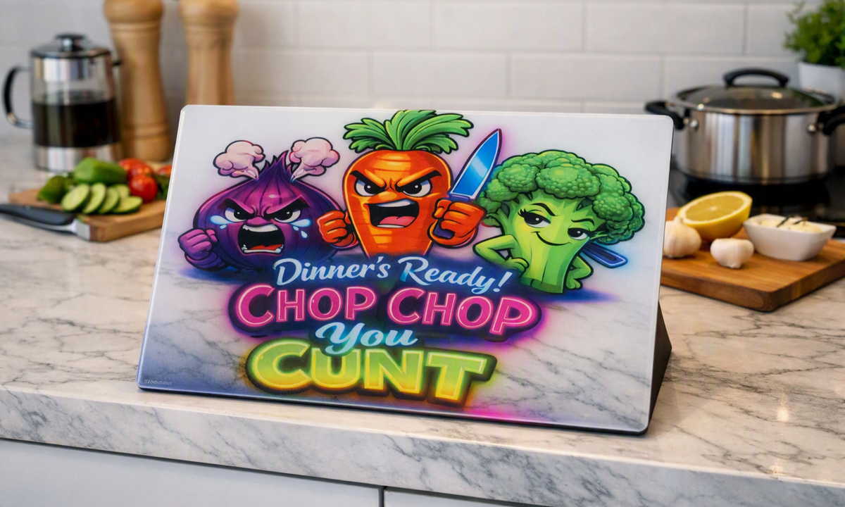 A glass cutting board sits on a worktop in a blurred kitchen setting. The cutting board displays a colourful image of an angry carrot holding a knife, an angry onion and a smug looking broccoli stem. Wording on the cutting board, in bright colourful lettering, reads Dinner's Ready! CHOP CHOP you CUNT