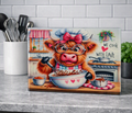 A rectangular glass chopping board standing on a kitchen counter, featuring a whimsical illustration of a fluffy brown cow in a chef hat and glasses mixing chocolate into a bowl. The sign reads 'WE COOK WITH LOVE' and is photographed against a real kitchen background with utensils, a tile wall, and a plant.