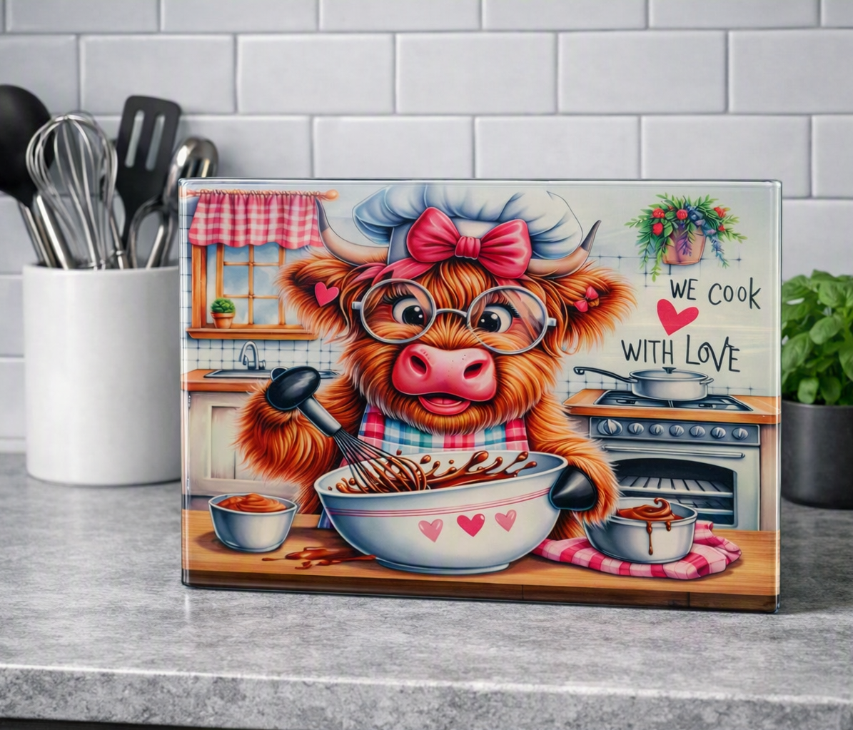 A rectangular glass chopping board standing on a kitchen counter, featuring a whimsical illustration of a fluffy brown cow in a chef hat and glasses mixing chocolate into a bowl. The sign reads 'WE COOK WITH LOVE' and is photographed against a real kitchen background with utensils, a tile wall, and a plant.