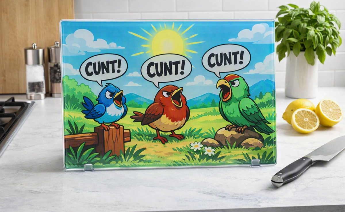 A rectangular glass cutting board, depicting 3 colourful birds, sat in a countryside setting, all saying the word CUNT! The cutting board sits on a marble kitchen worktop alongside a knife and freshly cut lemons