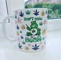 A white ceramic mug featuring a green Care Bear holding a cannabis joint, surrounded by colourful cannabis leaves and the text 'DON'T CARE BEAR'.