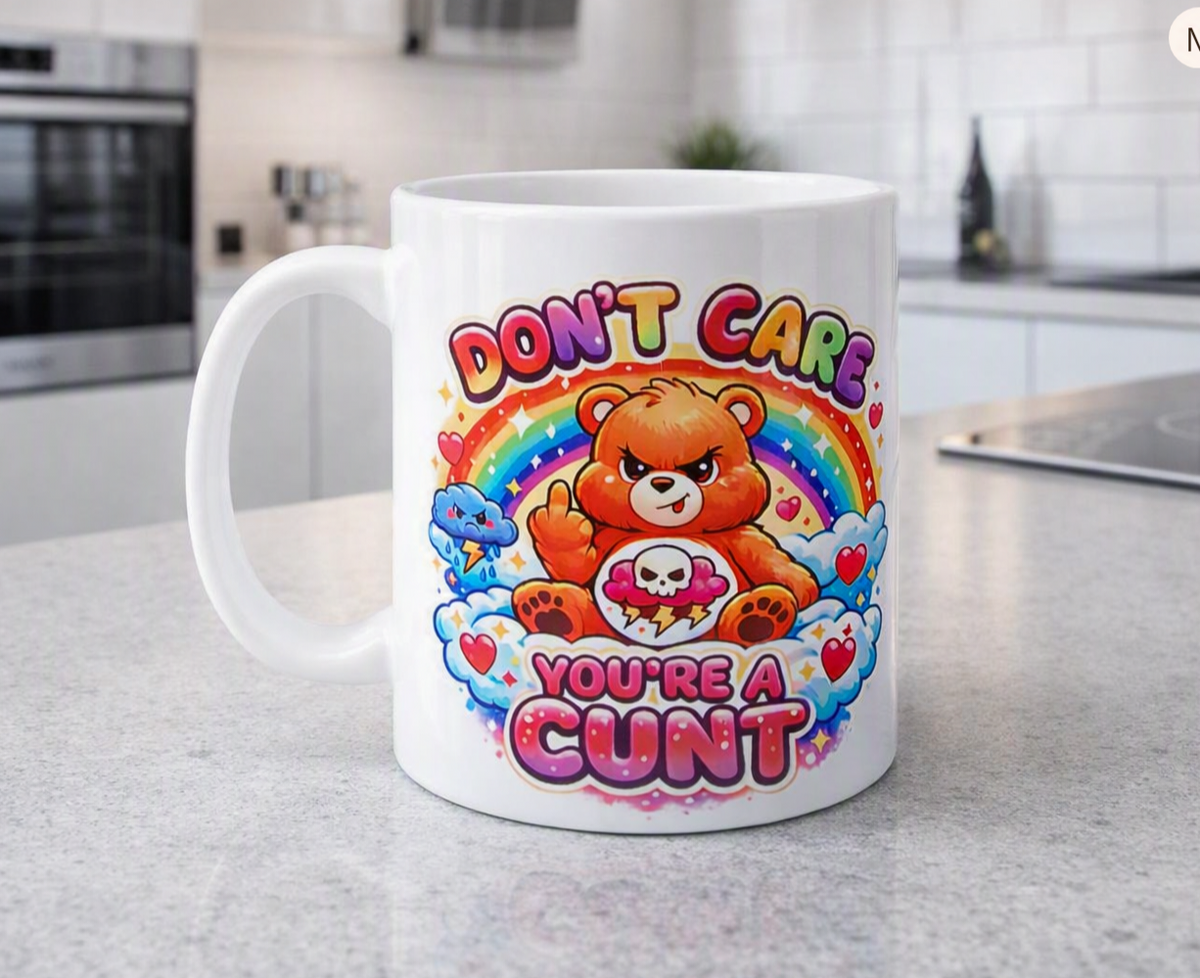 A white ceramic coffee mug with a colourful cartoon bear graphic and the text 'DON'T CARE YOU'RE A CUNT' sitting on a grey kitchen countertop.