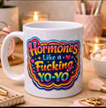 A white ceramic coffee mug with a colorful graphic that reads 'Hormones Like a Fucking YO-YO' in yellow, pink, blue, and orange lettering, set on a wooden desk with blurred cosmetic items and candles in the background.