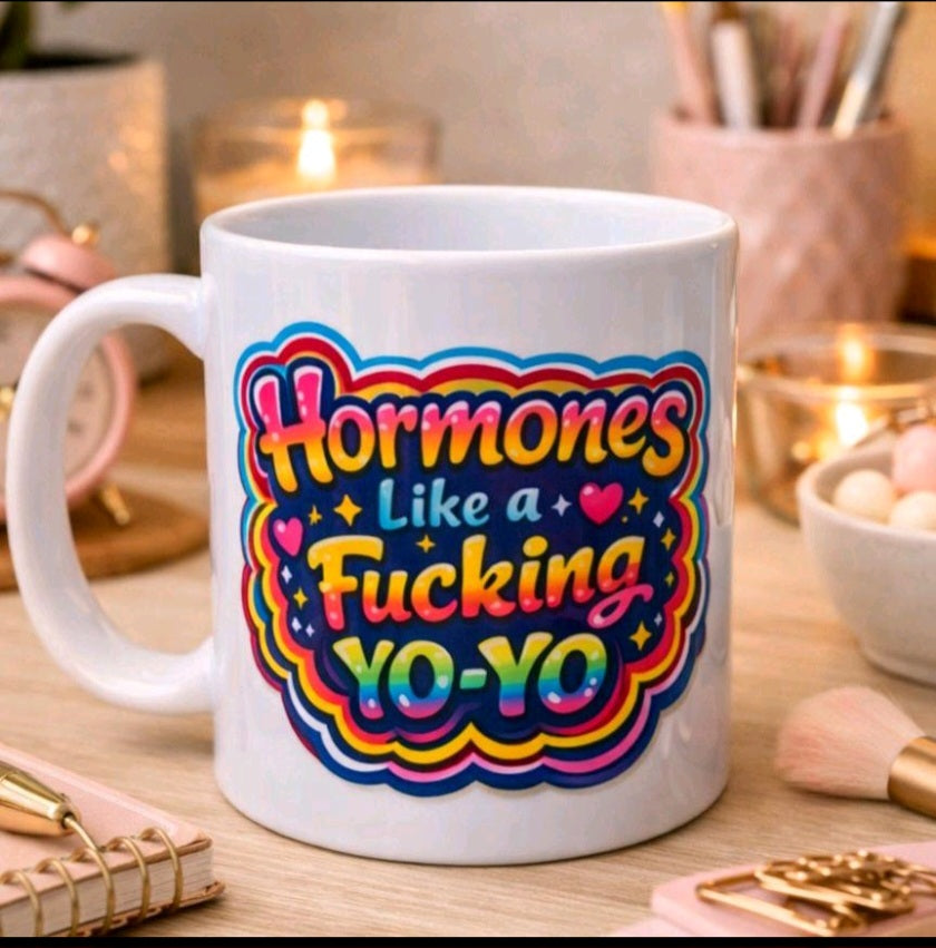 A white ceramic coffee mug with a colorful graphic that reads 'Hormones Like a Fucking YO-YO' in yellow, pink, blue, and orange lettering, set on a wooden desk with blurred cosmetic items and candles in the background.