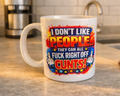 A white ceramic coffee mug with a bold, colorful graphic on a kitchen countertop. The graphic features the text 'I DON'T LIKE PEOPLE THEY CAN ALL FUCK RIGHT OFF CUNTS!' in red, yellow, and blue with rocket illustrations.