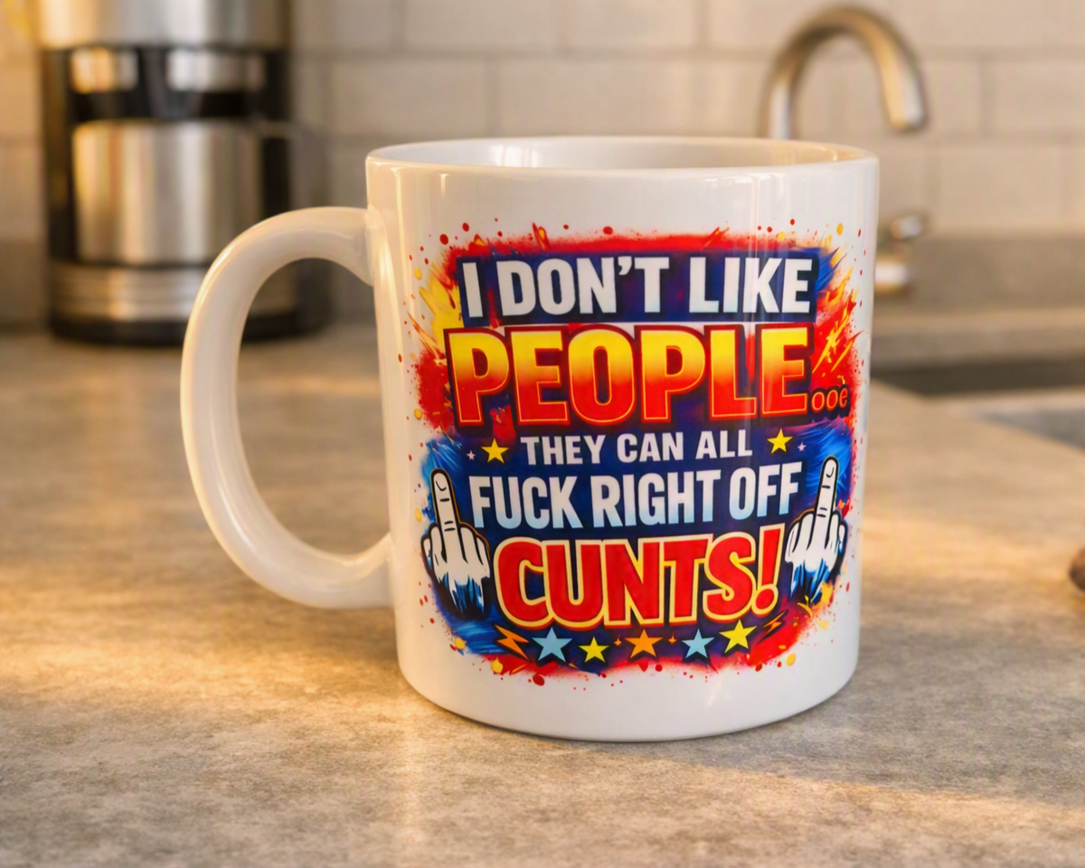 A white ceramic coffee mug with a bold, colorful graphic on a kitchen countertop. The graphic features the text 'I DON'T LIKE PEOPLE THEY CAN ALL FUCK RIGHT OFF CUNTS!' in red, yellow, and blue with rocket illustrations.