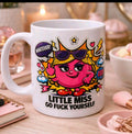 A white ceramic coffee mug with a colorful cartoon graphic of a Little Miss character wearing sunglasses, surrounded by speech bubbles and the text 'LITTLE MISS GO FUCK YOURSELF'. The mug is set on a wooden desk with blurred cosmetic and stationery items in the background.