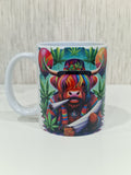 A white ceramic mug featuring a colorful graphic print of a highland cow wearing a rainbow-striped hat and scarf, surrounded by green cannabis leaves, set against a light wood background.