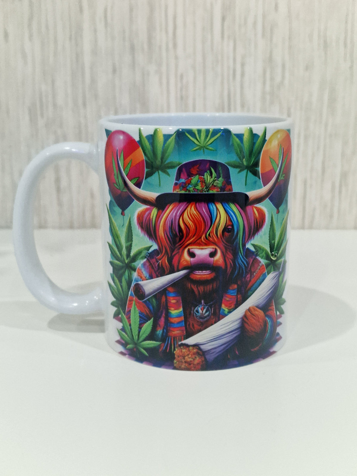 A white ceramic mug featuring a colorful graphic print of a highland cow wearing a rainbow-striped hat and scarf, surrounded by green cannabis leaves, set against a light wood background.