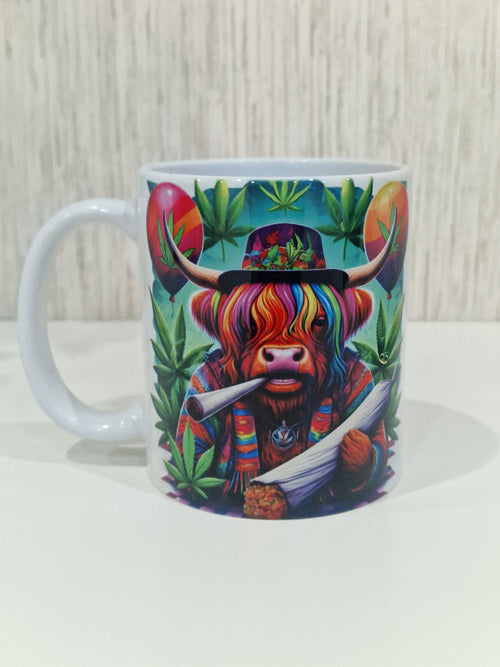 A white ceramic mug featuring a colorful graphic print of a highland cow wearing a rainbow-striped hat and scarf, surrounded by green cannabis leaves, set against a light wood background.