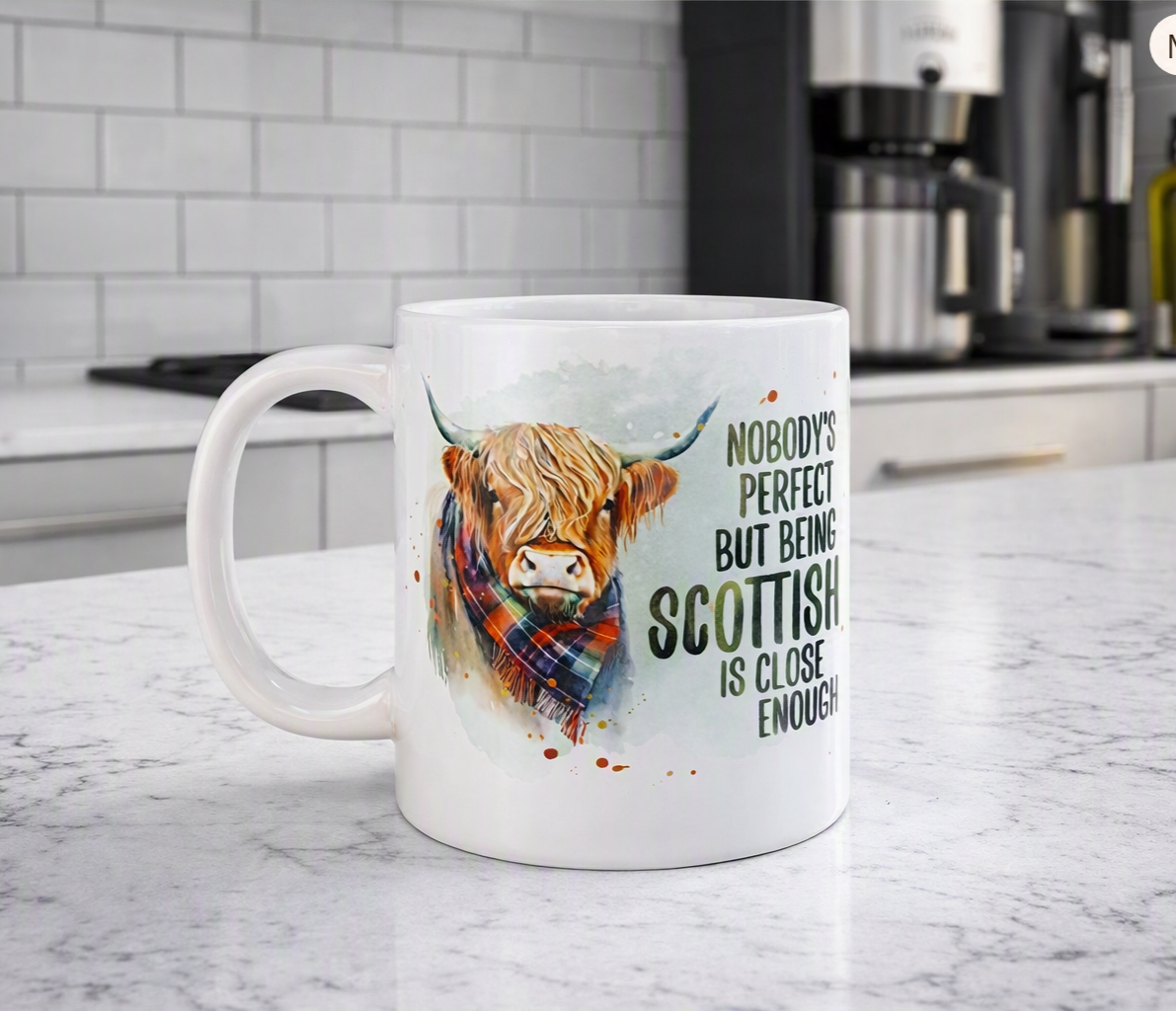 A white ceramic mug with a watercolor illustration of a Highland cow in a plaid scarf and the text 'NOBODY'S PERFECT BUT BEING SCOTTISH IS CLOSE ENOUGH!' sits on a marble kitchen countertop with a blurred kitchen background.