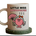 A white ceramic coffee mug with the text 'LITTLE MISS MENOPAUSE' and a cartoon illustration of a pink character holding a fan, sitting on a round green coaster against a white background.
