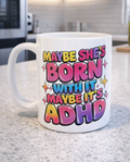 A white ceramic coffee mug with a colorful text graphic reading 'MAYBE SHE'S BORN WITH IT MAYBE IT'S ADHD' in pink, yellow, blue, and purple bubble letters, sitting on a speckled kitchen countertop.