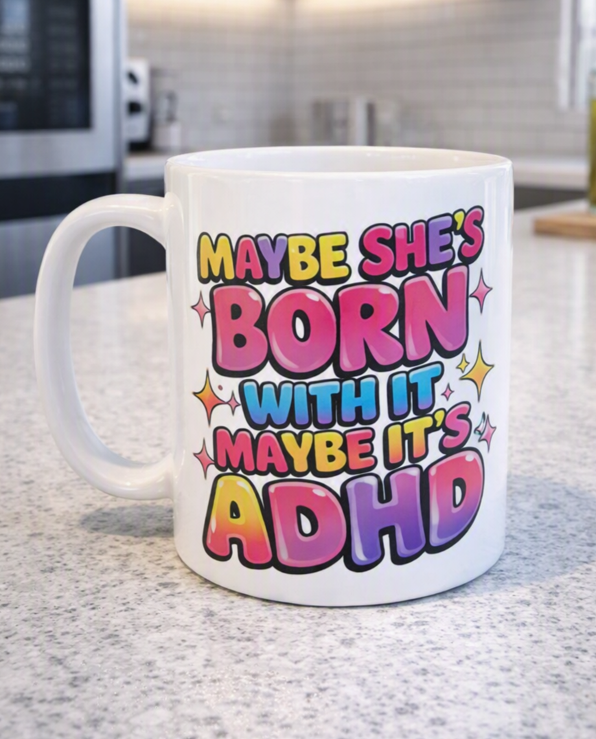 A white ceramic coffee mug with a colorful text graphic reading 'MAYBE SHE'S BORN WITH IT MAYBE IT'S ADHD' in pink, yellow, blue, and purple bubble letters, sitting on a speckled kitchen countertop.