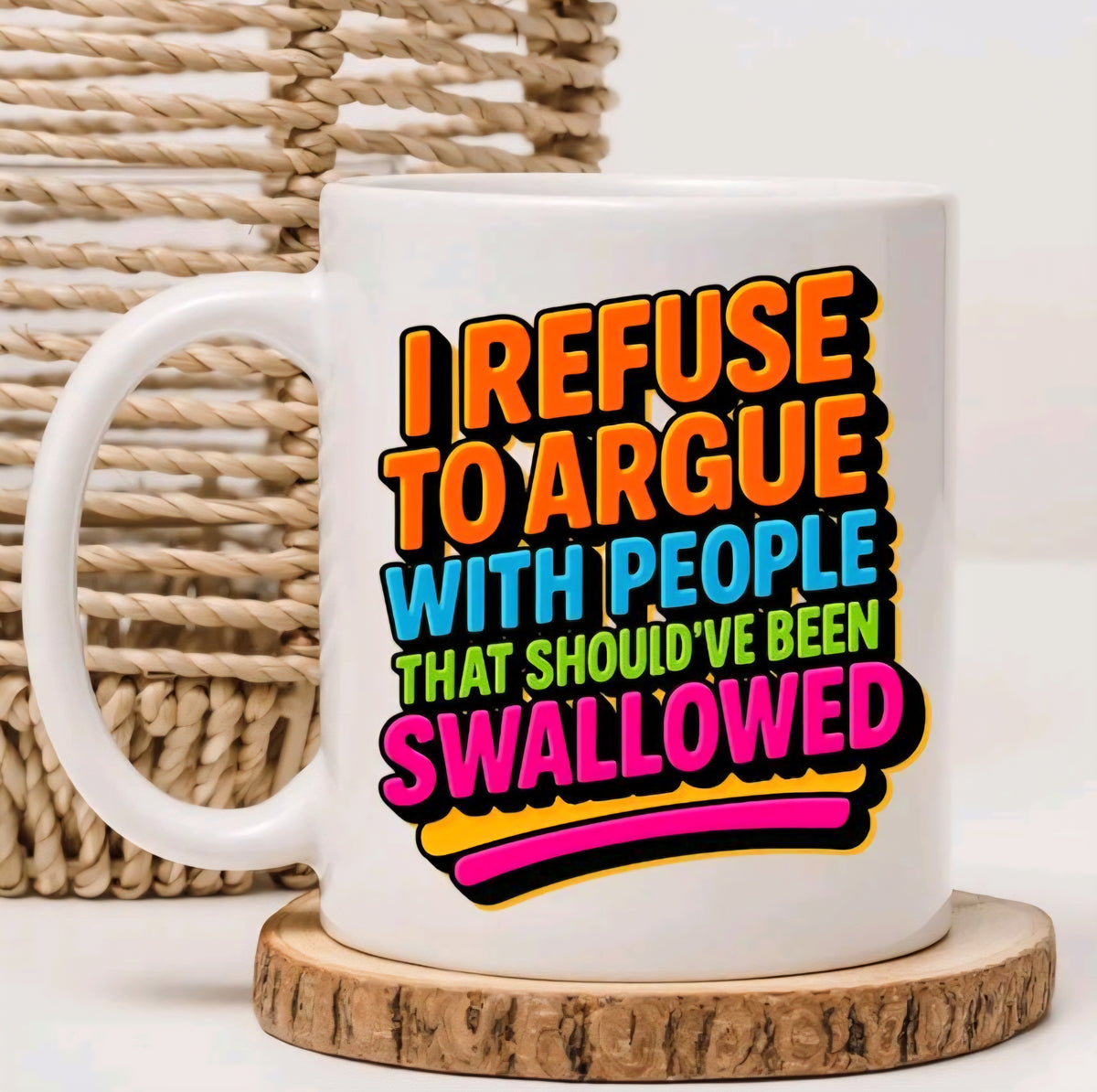 A white ceramic mug with a colorful text graphic reading 'I REFUSE TO ARGUE WITH PEOPLE THAT SHOULD'VE BEEN SWALLOWED' in orange, blue, green, and pink lettering, styled on a wooden coaster with a woven basket in the background.