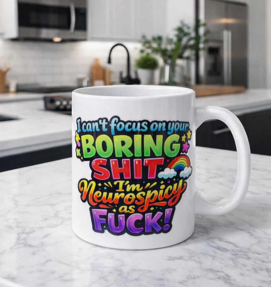 A white ceramic coffee mug with a colorful, bold graphic text design reading 'I can't focus on your BORING SHIT I'm Neurospicy as FUCK!' sits on a white marble countertop with a blurred modern kitchen in the background.