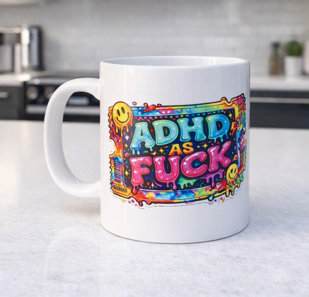 A white ceramic mug with a colorful graphic that reads 'ADHD AS FUCK' in bold letters, decorated with yellow smiley faces and paint splatter details, sitting on a light gray countertop with a blurred kitchen background.