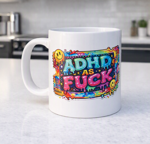 A white ceramic mug with a colorful graphic that reads 'ADHD AS FUCK' in bold letters, decorated with yellow smiley faces and paint splatter details, sitting on a light gray countertop with a blurred kitchen background.