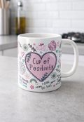 A white ceramic coffee mug with a pink heart and floral doodle design and the words Cup of Positivity, sitting on a grey countertop in a blurred kitchen setting.