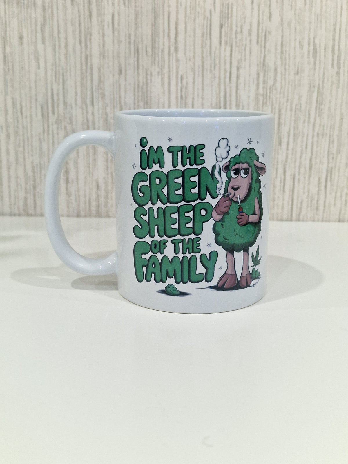 A white ceramic mug featuring a green cartoon sheep character and the text 'I'M THE GREEN SHEEP OF THE FAMILY' in green lettering, set against a light wood-grained background.
