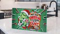 A rectangular green Christmas sign featuring the Grinch in a Santa hat with the text 'MAY YOUR CHRISTMAS BE AS MERRY AS A CUNT' and candy cane illustrations, displayed on a kitchen countertop.