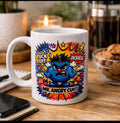 A white ceramic coffee mug with a colorful graphic of an angry blue cartoon character surrounded by comic-style bursts and text reading 'FUCK OFF', 'DICKHEAD', and 'MR. ANGRY CUNT'.