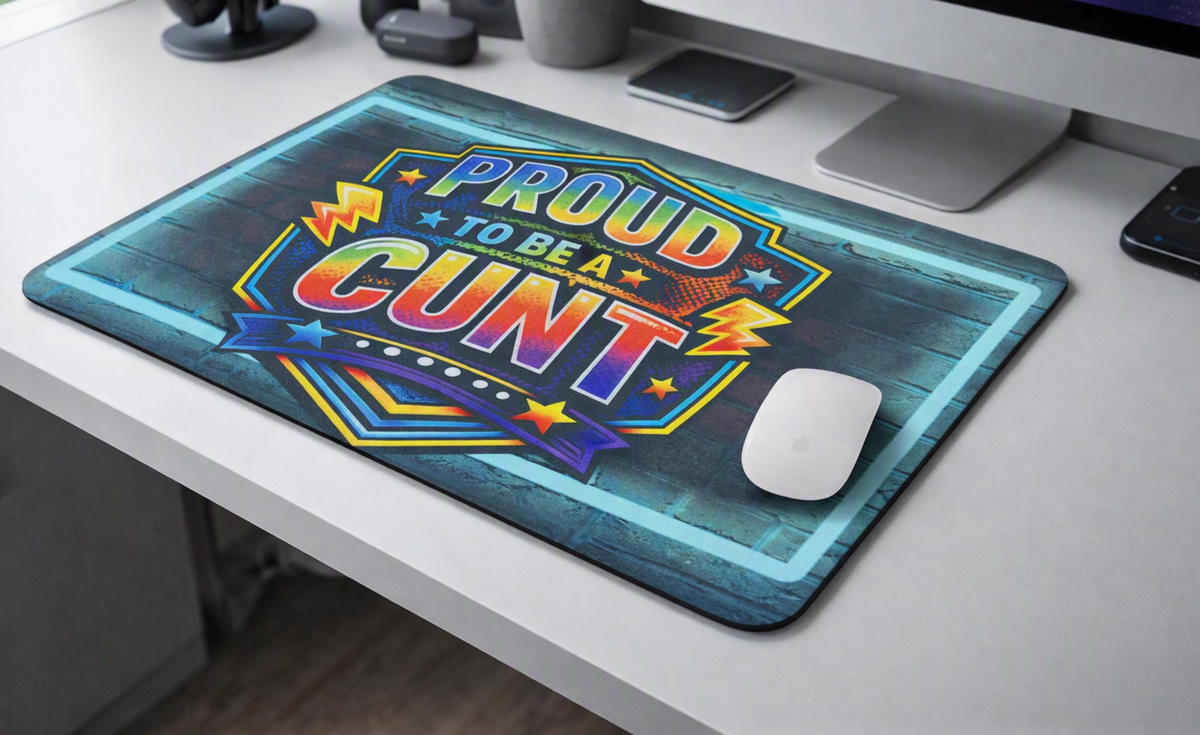 A rectangular mouse mat with a colourful graphic reading 'PROUD TO BE A CUNT' sits on a white desk next to a white computer mouse. The mat has a blue/grey brick effect background with a bright multicolor text design and decorative border.