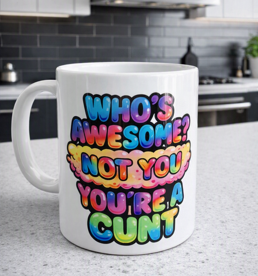 A white ceramic coffee mug with a colorful rainbow gradient text design that reads 'WHO'S AWESOME? NOT YOU YOU'RE A CUNT', set on a kitchen countertop with a blurred kitchen background.